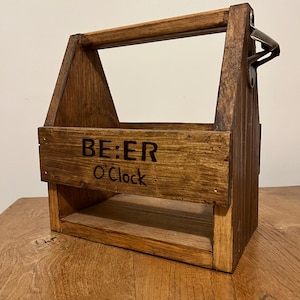 Puede incluir: Un portabotellas de cerveza de madera con asa y abridor de botellas integrado. La parte delantera del portabotellas tiene las palabras "BE:ER O'Clock" en letras negras. El portabotellas está hecho de madera teñida y tiene un aspecto rústico.