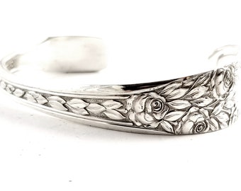 May include: A silver bracelet with a floral design featuring roses and leaves. The bracelet is made from a single piece of metal that is bent into a circle.