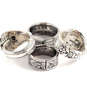 May include: Four silver spoon rings with engraved messages. The rings are stacked on top of each other. The messages on the rings are "I am enough", "Love Mom", "Have a life less ordinary with me", and a skull and crossbones.