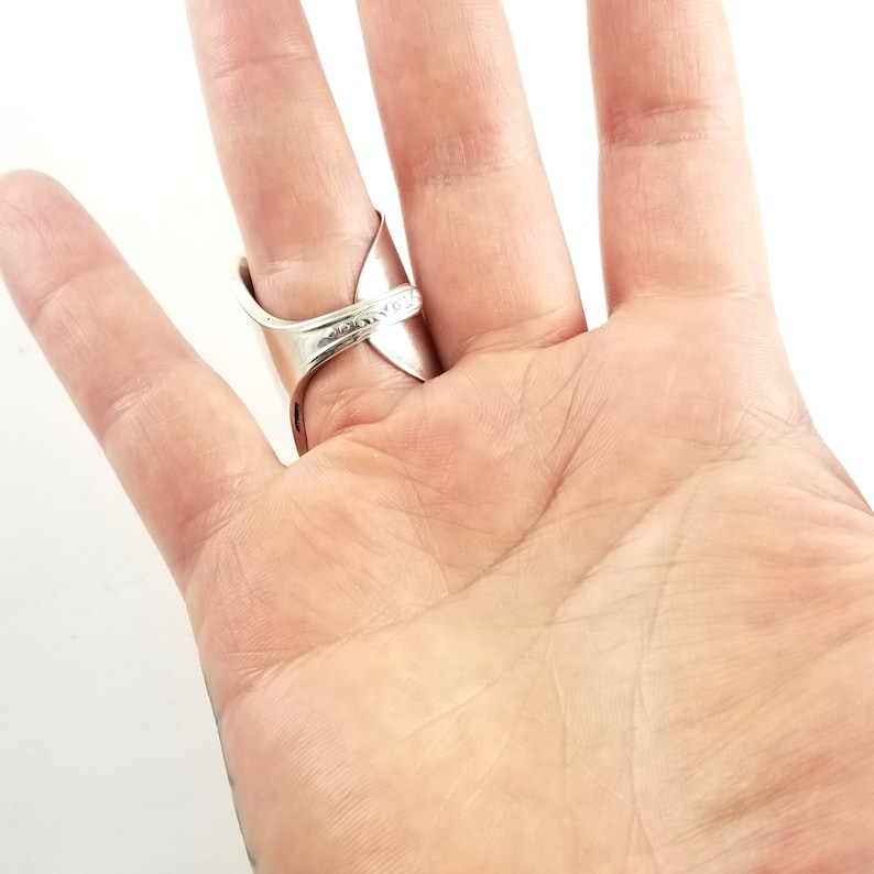 May include: A silver ring made from a bent spoon. The ring is worn on the index finger of a hand.
