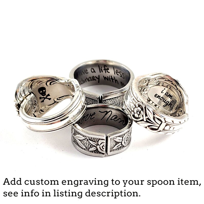 May include: Four silver spoon rings with different engraved messages. The rings are stacked on top of each other. The messages are "live a life less ordinary with me", "love Mom", "i am enough", and a skull and crossbones.