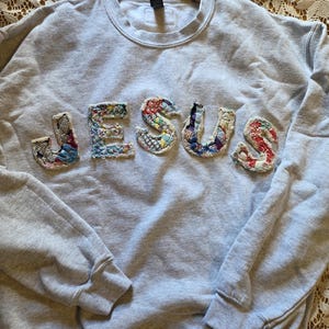 May include: A light gray crewneck sweatshirt featuring the word JESUS appliqued across the chest. The letters are constructed from various colorful, patterned fabric scraps with frayed edges, creating a textured, multi-colored appearance.