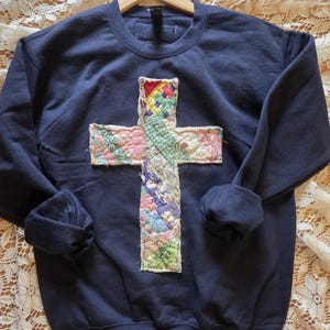 May include: A navy blue sweatshirt with a patchwork cross design. The cross is made of various fabric patterns in shades of pink, green, blue, and yellow. The sweatshirt has a crew neck and long sleeves.