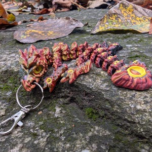 Puede incluir: Un llavero de dragón rojo y amarillo con un anillo de llave plateado. El dragón está hecho de plástico y tiene un diseño detallado.