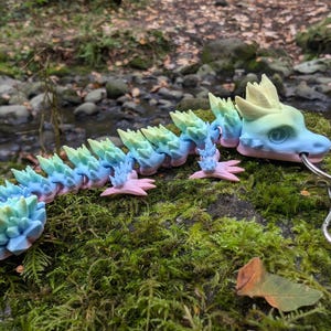 Op de afbeelding: Een regenboogkleurige, gearticuleerde draken sleutelhanger met een zilveren sleutelring. De draak is gemaakt van plastic en heeft een stekelig, kristallijn ontwerp. De draak is geposeerd met zijn hoofd gedraaid naar de zijkant en zijn mond lichtjes open.