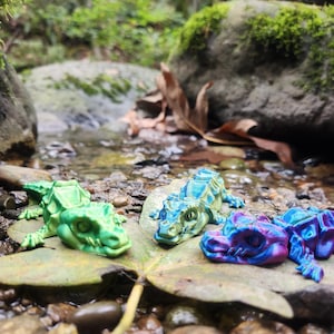 May include: Three small, colourful, plastic alligator figurines on a leaf near a stream. The alligators are green, blue and purple with iridescent paint.