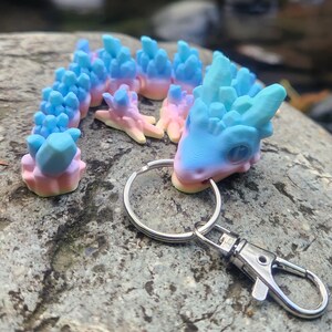 May include: A pastel blue and pink crystal dragon keychain with a silver key ring and clasp. The dragon is made of a 3D printed material and has a detailed design.