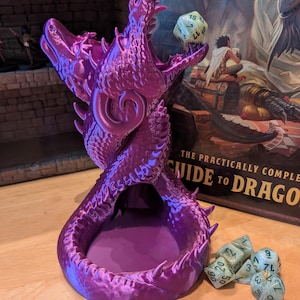 May include: Purple 3D printed dragon dice tower with a green and gold dice set. The dice tower is shaped like a dragon with three heads and a large opening in the centre. The dice are scattered around the base of the tower.