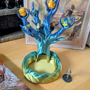 May include: A blue and green 3D printed tree with branches that hold dice. The tree has a base that is a dice rolling tray. The tree is decorated with a variety of dice in different colours.
