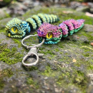 Puede incluir: Un llavero de serpiente iridiscente de colores con un anillo y un cierre plateados. La serpiente está hecha de plástico y tiene una superficie texturizada. La serpiente es un diseño popular para llaveros y es una excelente manera de agregar un toque de personalidad a tus llaves.