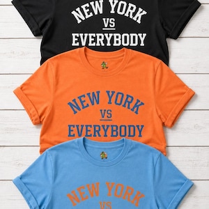 May include: Three short-sleeved t-shirts stacked vertically. The top shirt is black with white text, the middle is orange with blue text, and the bottom is light blue with orange text. Each shirt features the text NEW YORK VS EVERYBODY.