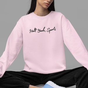 Könnte beinhalten: Ein rosa Sweatshirt mit dem Text "Hell Yeah, Sports" in schwarzer Kursivschrift.