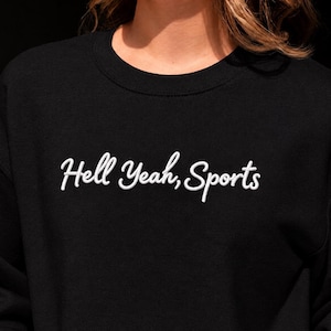 May include: A black crewneck sweatshirt featuring the white embroidered text Hell Yeah, Sports in a cursive script font across the chest. The garment has long sleeves and a ribbed neckline, providing a simple and clean look for casual wear.