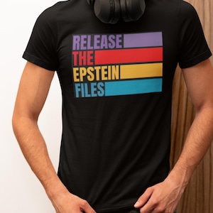 Puede incluir: Camiseta negra con el texto "RELEASE THE EPSTEIN FILES" en un diseño rectangular colorido. El texto es de color morado, rojo, amarillo y azul. La camiseta es de cuello redondo.