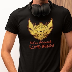 Puede incluir: Una camiseta negra de manga corta con un gráfico dorado y rojo de una cabeza robótica con rasgos afilados y angulosos. Debajo del gráfico, el texto "We've Achieved SOMETHING!" aparece en una fuente roja de estilo manuscrito.