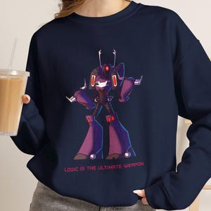 May include: A navy blue crewneck sweatshirt featuring a graphic of a purple and black robotic character with a single red eye. Below the character, the text reads "LOGIC IS THE ULTIMATE WEAPON" in red capital letters. The design includes a small purple emblem on the sleeve.