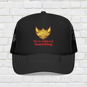 May include: Black trucker hat with a gold and red emblem and the text "We've Achieved Something." The hat has a mesh back and a curved brim. The emblem is a stylized design.