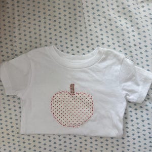 May include: White short-sleeved t-shirt with an apple applique. The apple is made of white fabric with red polka dots and a red stitched outline. The stem is a brown, plaid fabric.