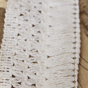 May include: White crocheted trim with a fringe detail. The trim is made of a soft, delicate yarn and has a delicate, lacy pattern.