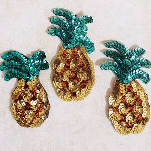 May include: Three gold and green pineapple-shaped patches made of sequins. The pineapples have a gold body and green leaves.