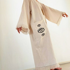 May include: A long, off-white cotton robe with fringe detailing. The robe has a large pocket on the left side with an embroidered eye design.