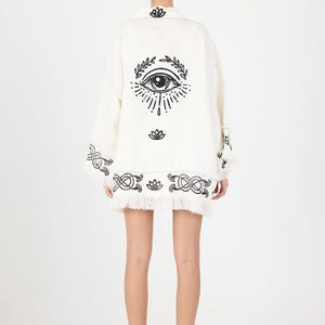 Puede incluir: Conjunto de chaqueta y falda blancos con diseños bordados en negro de ojos y flores. La chaqueta tiene mangas largas y un dobladillo con flecos. La falda también tiene un dobladillo con flecos. El conjunto se lleva con sandalias negras.