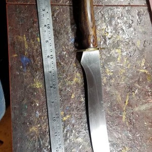 May include: A long, curved knife with a brown wooden handle and a silver blade. The blade is about 12 inches long and the handle is about 5 inches long. The knife is resting on a wooden surface.