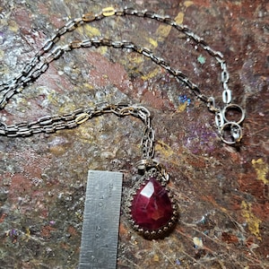May include: A silver chain necklace with a teardrop-shaped pendant featuring a large, faceted, red gemstone set in a silver filigree frame.
