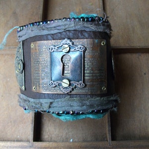 May include: A brown leather bracelet with a metal keyhole charm and a quote about the power of imagination. The bracelet is decorated with turquoise and blue beads and fabric.