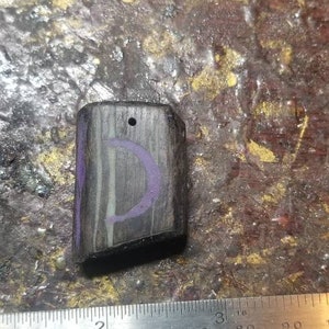May include: A small, dark wood pendant with a purple crescent moon design. The pendant has a hole at the top for a cord or chain.