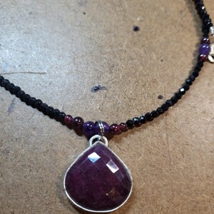May include: A purple gemstone pendant necklace with a teardrop-shaped stone set in silver. The necklace is made with black beads and small purple and red beads.