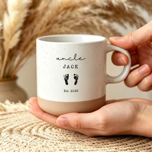 May include: A white ceramic mug with a speckled finish and a tan base. The front features the black text "uncle JACK" above two small black baby footprints and the text "Est. 2026". The mug has a curved handle on the right side.