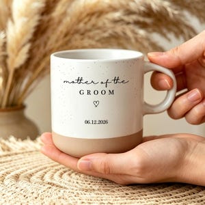 Op de afbeelding: Een witte keramische mok met een gespikkelde afwerking en een beige basis. De tekst "mother of the GROOM" is in het zwart gedrukt, gevolgd door een klein harticoontje en de datum "06.12.2026". De mok heeft een gebogen handvat en een cilindrisch lichaam.