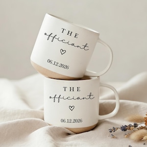 Puede incluir: Dos tazas de cerámica blanca apiladas. Cada taza tiene las palabras "THE officiant" en fuente manuscrita, un símbolo de corazón y la fecha "06.12.2026". La base de las tazas es de color beige. Flores secas en el fondo.