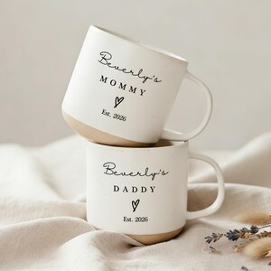 Puede incluir: Dos tazas de cerámica blanca apiladas. La taza superior dice "Beverly's MOMMY" con un corazón y "Est. 2026". La taza inferior dice "Beverly's DADDY" con un corazón y "Est. 2026". La base de cada taza es de color beige.