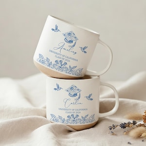 Puede incluir: Dos tazas de cerámica blanca apiladas. Cada taza tiene una base marrón claro y un cuerpo blanco con diseños florales y colibríes azules. El texto en las tazas dice "Amelia" y "Carlin", "University of California Class of 2026".