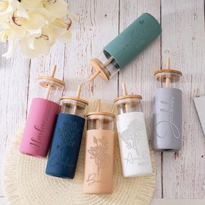 May include: A collection of glass tumblers with bamboo lids and straws. Each tumbler has a colored silicone sleeve with a floral design and a name or title. Colors include pink, blue, beige, white, and gray.