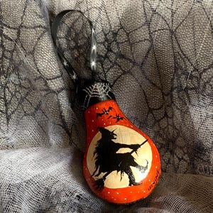 May include: Hand-painted Halloween ornament shaped like a lightbulb. The ornament is red with a white moon and a black silhouette of a witch flying on a broom. Black bats fly across the top. A black ribbon with white spiders is attached.