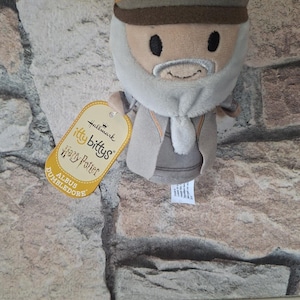 Könnte beinhalten: Ein Plüschtier der Figur Albus Dumbledore aus der Hallmark Itty Bittys Harry Potter Kollektion. Die Figur trägt einen beigen Hut mit goldener Verzierung, einen grauen Bart und ein beiges Gewand. Ein gelbes Etikett zeigt die Aufschrift Hallmark Itty Bittys.