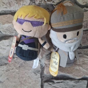 Op de afbeelding: Twee pluche sleutelhangers met een blonde figuur met paarse oogbescherming en een donker pak, en een figuur met een grijze baard, grijze hoed en gewaad. Beide speeltjes hebben Hallmark-labels, waarvan er één is gelabeld als Itty Bittys Harry Potter Albus Dumbledore.