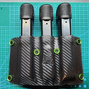 Pode incluir: Um coldre de couro preto com três slots para carregar carregadores. O coldre tem um padrão de fibra de carbono e parafusos verdes.