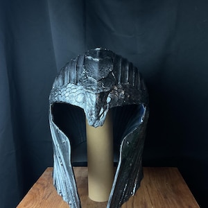 以下が含まれることがあります： ヘビのようなデザインの濃い灰色のファンタジーヘルメット。ヘルメットは、テクスチャ加工された表面、眉毛部分の精巧なヘビの頭、フレア状の頬ガードが特徴です。ヘルメットは茶色の段ボールチューブに展示されています。