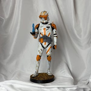 May include: A detailed figurine of a Star Wars clone trooper in white armor with orange accents. The figure is holding a blue object and stands on a circular base. The helmet and armor show signs of wear.