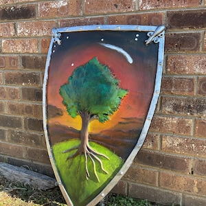 May include: Hand-painted shield featuring a tree with a green canopy and exposed roots on a green hill. The background depicts a sunset with a shooting star and a night sky. The shield has a silver metal trim.