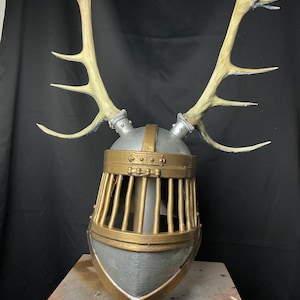 May include: A medieval-style helmet with a cage-like face guard and a pair of large, light-colored antlers. The helmet is primarily gray with gold-colored accents. The antlers are attached to the top of the helmet.