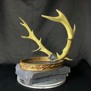May include: A gold-colored crown with an antler design, set atop a gray, layered base. The crown features intricate detailing and is mounted on a circular platform. The antlers extend upwards, creating a unique decorative piece.
