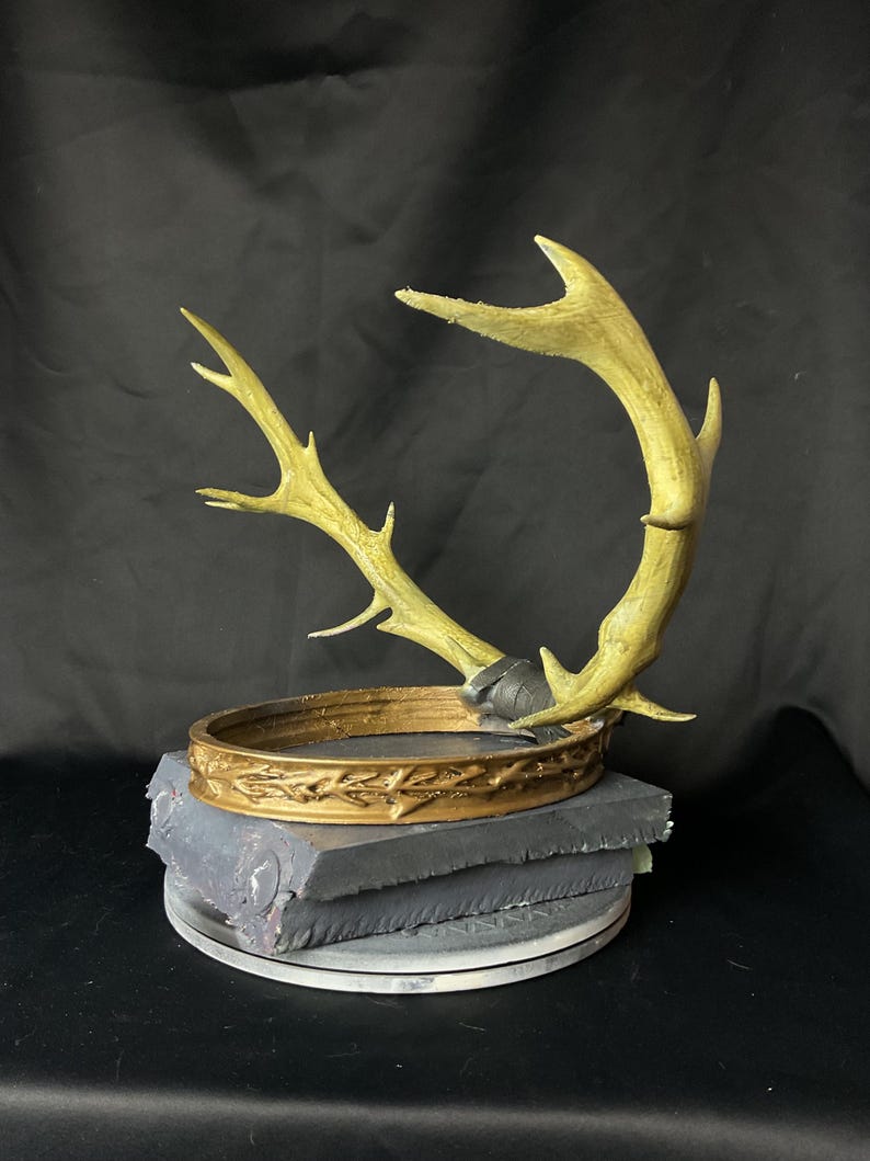 May include: A gold-colored crown with an antler design, set atop a gray, layered base. The crown features intricate detailing and is mounted on a circular platform. The antlers extend upwards, creating a unique decorative piece.