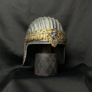 May include: A silver and gold helmet with a decorative design. The helmet has a ridged top and a gold band with a detailed pattern. A small, ornate figure is centered on the front.