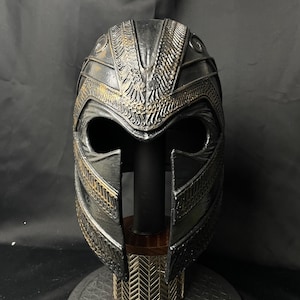 以下が含まれることがあります： 精巧なディテールが施された黒と金のヘルメット。ヘルメットは、質感のある表面と、額と側面に装飾的な模様が特徴です。金色の模様のベースが付いたスタンドに展示されています。