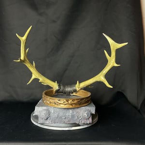 May include: A golden-colored antler crown with a decorative band and a gray base. The crown features a detailed design and is likely a costume piece or decorative item. The antlers are attached to the band with a dark material.